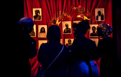 Makeshift Gatherings 2019 - Image by Bryony Jackson - Four people are in a dark room viewing a wall with a red curtain and multiple photos hanging on it.