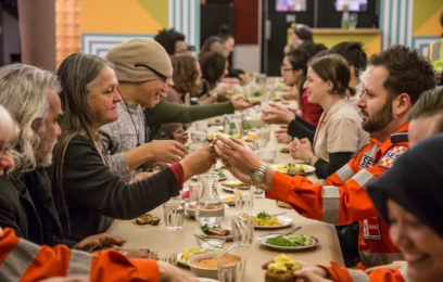 Refuge - A group of people sit around a long dining table. They are passing objects to each other across the table.