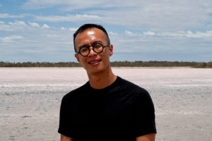  A man with an inland pink lake behind him smiling with round black glasses and short black hair