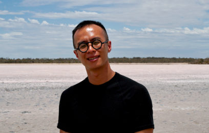 A man with an inland pink lake behind him smiling with round black glasses and short black hair