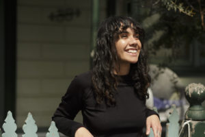 a woman with long black curly hair leans on a picket fence with a huge smile on her face