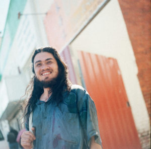 image of a man with long dark hair and a green shirt smiling against an orange background