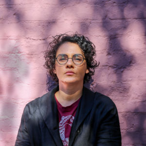 a person with short curly hair and freckles against a pale purple background with black rimmed glasses looking slightly down at the camera