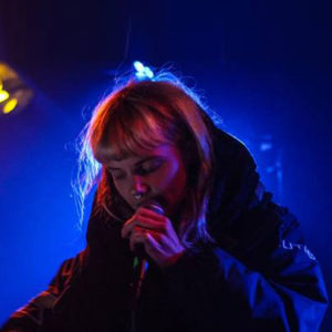 a figure in a dark space speaking into a microphone with a blue background