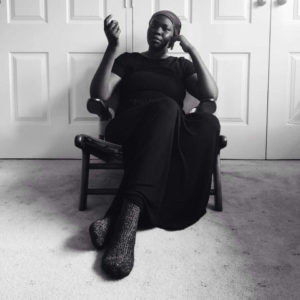 black and white image of the artist who is seated with black hair covering and holding one hand up in front of cupboard doors