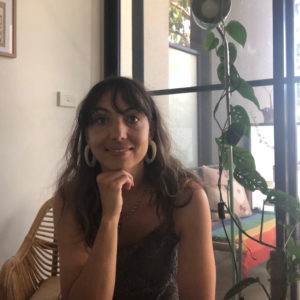 a woman resting her chin on her hand with a green grey top on and houseplants behind