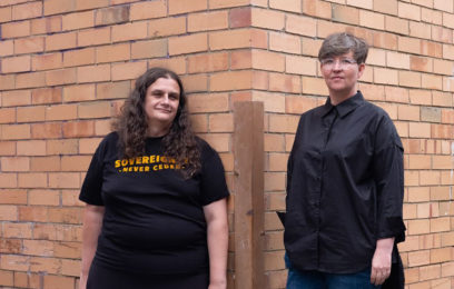 First Assembly - Two people stand against a brick wall. One has long curly brown hair and is wearing a t-shirt, the other has short grey hair with glasses and is wearing a black collared shirt.
