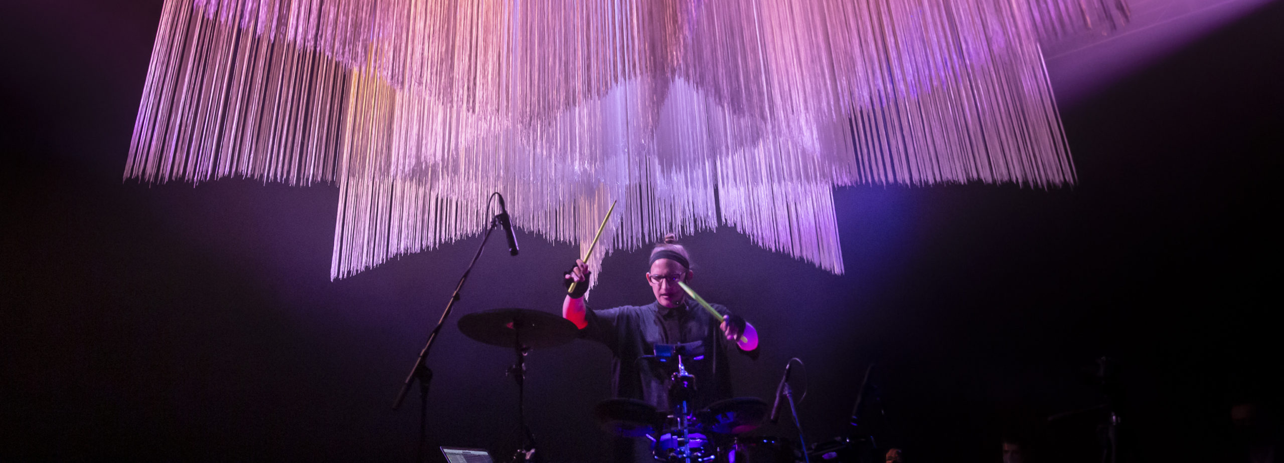 SINK - Simona Castricum X Carla Zimbler by Bryony Jackson - A woman plays the drums on stage. There is a geometric installation made from long strands of fringing suspended above her head and it is lit by pink, purple, and orange lights. She is wearing glasses, black fingerless gloves, a black shirt, and a black headband with her hair in a bun.