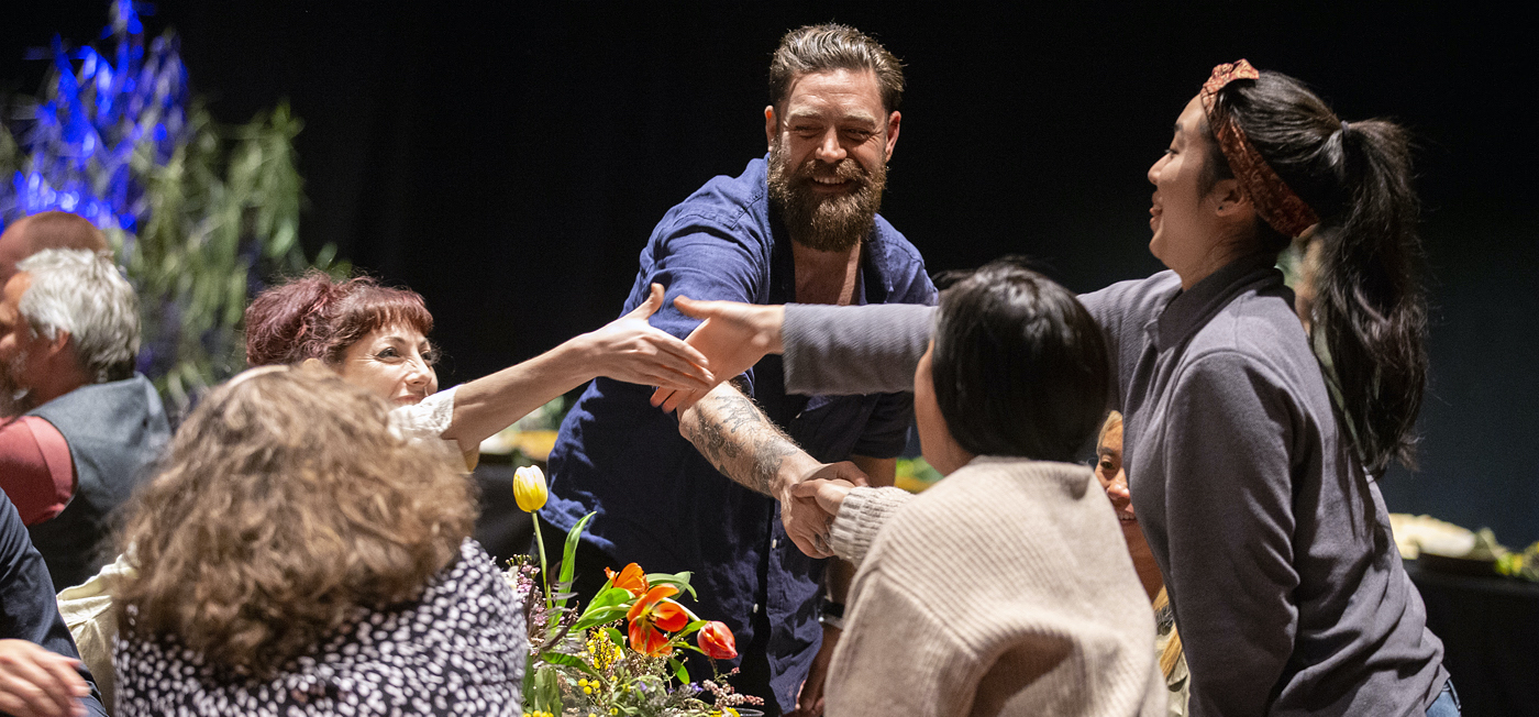 Fail Better: with Chris Cheers Arts House Refuge 2019 Displacement Words Nourish Neighbours_image Harjono Djoyobisono - A group of five people are sitting around a table, they are reaching their hands into the middle to hold hands. There are orange flowers in the middle of the table.