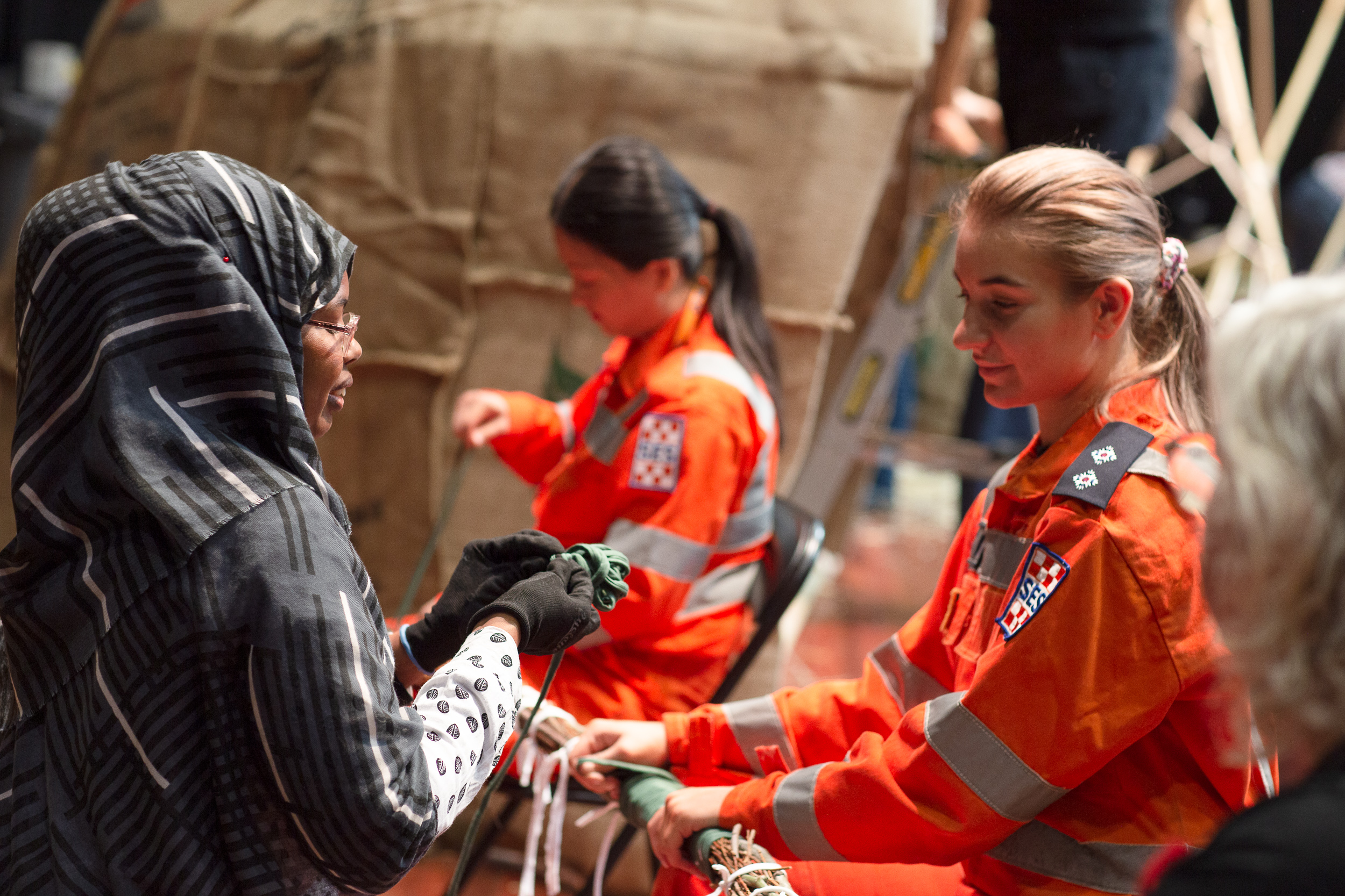 Refuge 2021 Documentary - Portage: Shelter2Camp by Jen Rae, photo by Bryony Jackson - Two emergency services people wearing high-vis orange uniforms. One with a blond ponytail is holding a rope structure with a woman in a black headscarf