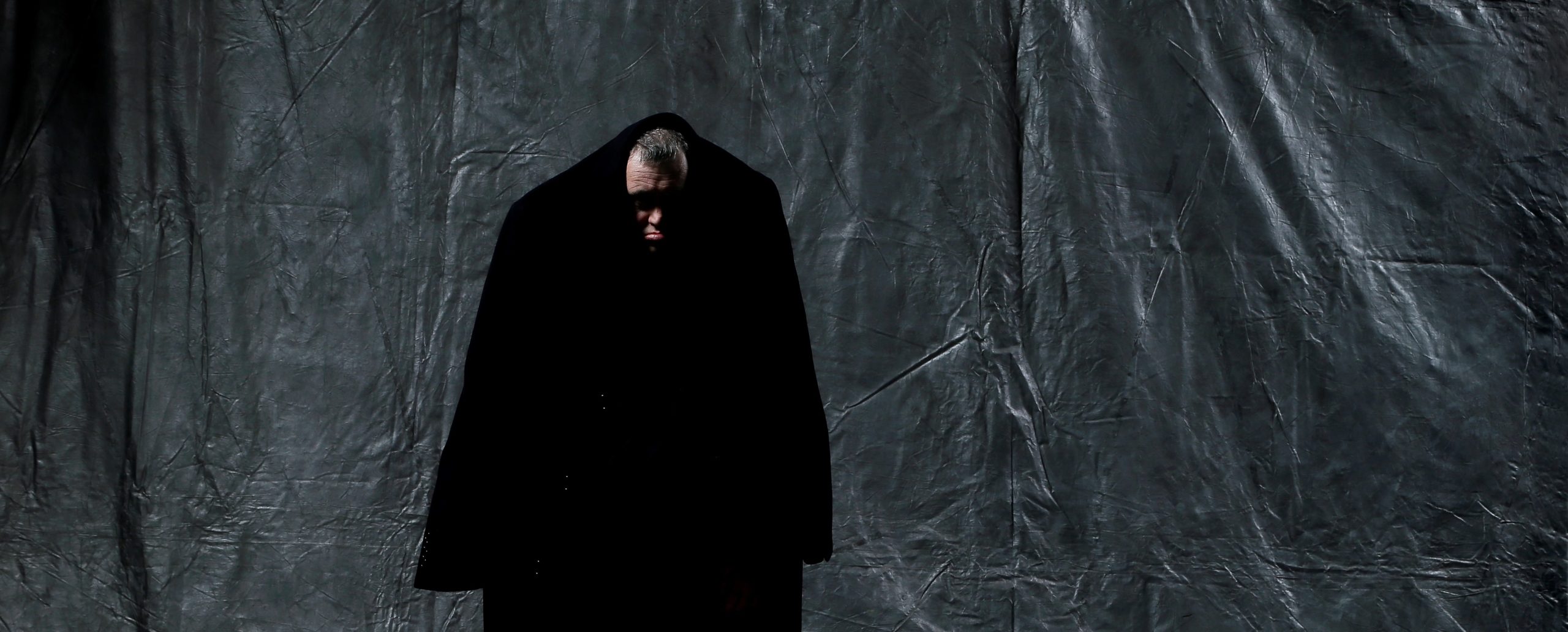 Entity - Back 2 Back Theatre - A man with curly grey hair on top of his head stands against a background of wrinkled dark grey fabric. He is barefoot and is wearing dark pants and an oversized coat. The coat is pulled up over his head and he has a sad expression on his face.