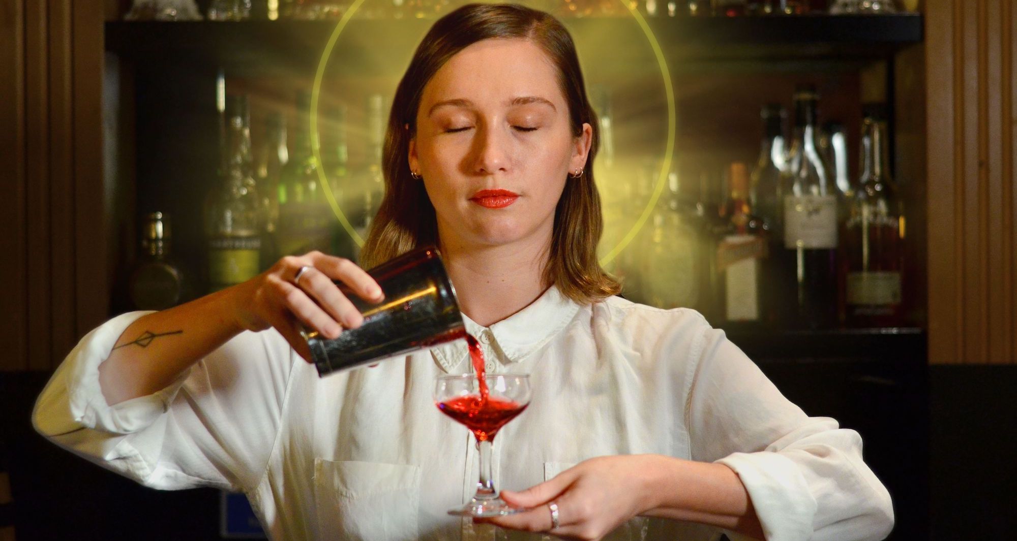 Love Me (Bar) Tender - Stage Mom - Photo by Peter Frith - A woman in a white shirt stands in front of a bar with her eyes closed. She has a gold halo imposed behind her head and she is pouring a red drink from a silver shaker into a small coupe glass in her left hand. She has shoulder length brown hair, red lipstick, and a serene expression on her face.