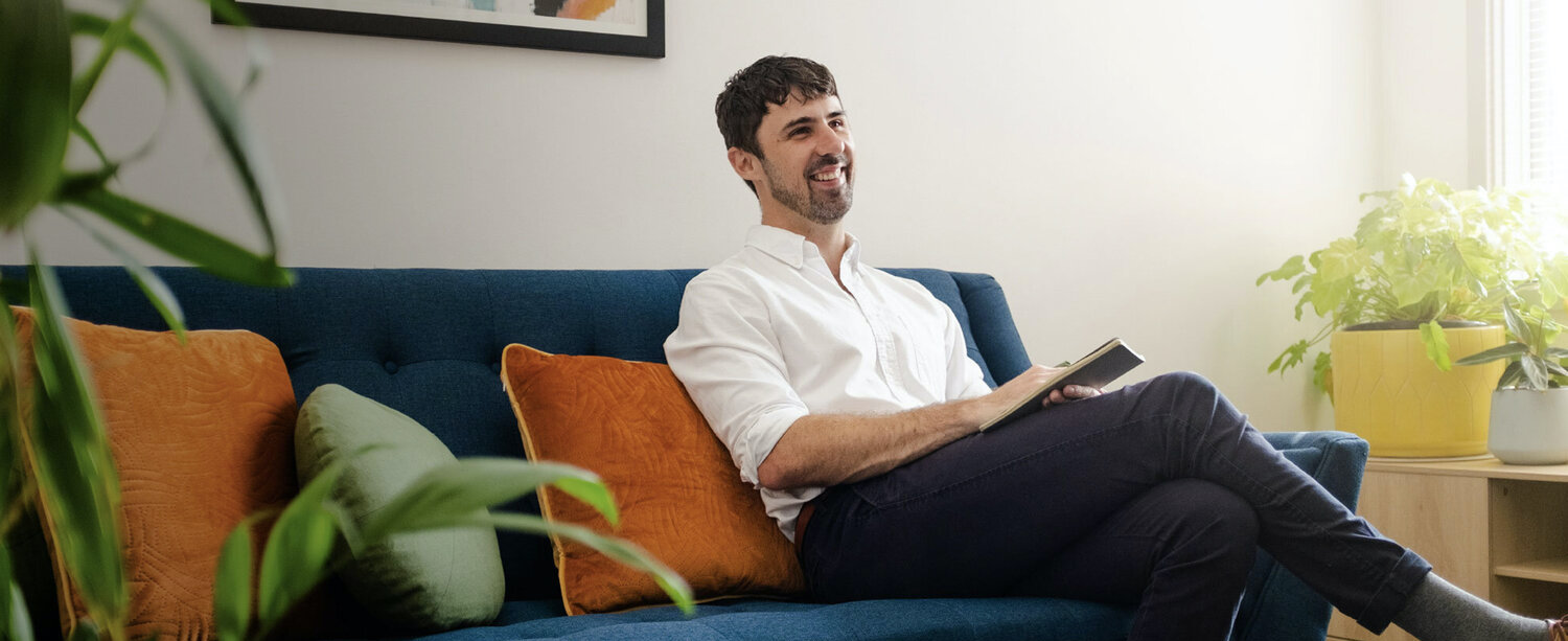 Chris Cheers - Fail Better - A man with short dark hair and dark stubble sits on a blue couch. He is holding a book. There are orange cushions on the couch and a plant in a yellow pot in the background. The man is smiling.