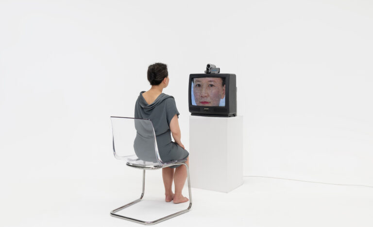 Photo of woman in a chair, facing a video camera and TV that shows her face.